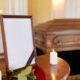 A small table displays a picture frame, lit white candles, and a rose in front of a wooden casket at a funeral.