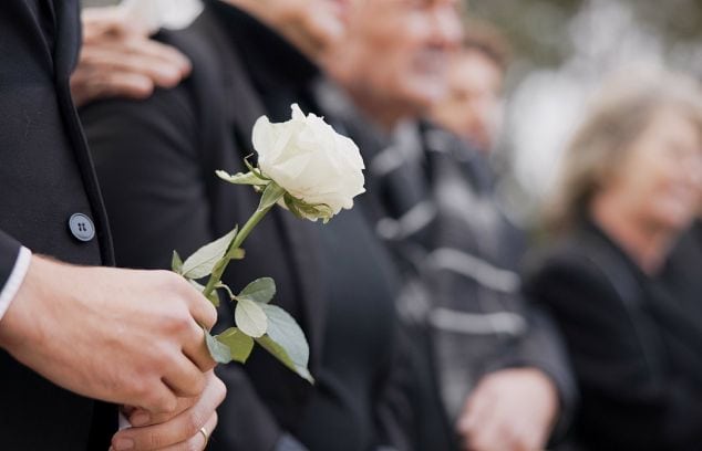 A line of people standing and wearing dark clothes at a memorial service. A man holds a white rose in the foreground.