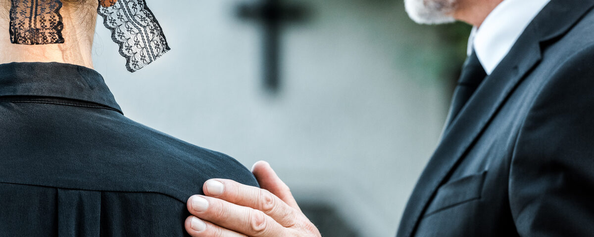 Two funeral guests face away from view. The older man places his hand on the woman's shoulder next to him.