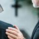 Two funeral guests face away from view. The older man places his hand on the woman's shoulder next to him.