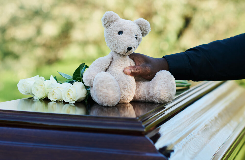 A man's arm in a black, long-sleeve shirt places an off-white teddy bear on a casket next to four white roses.